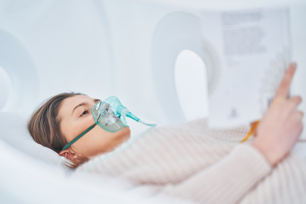 A person resting inside a hyperbaric oxygen therapy chamber wearing a clear oxygen mask