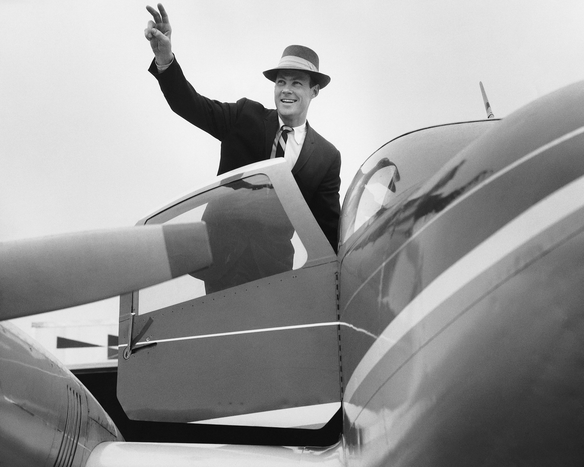 A confident traveler in a suit and hat waving from the cockpit door of a vintage aircraft