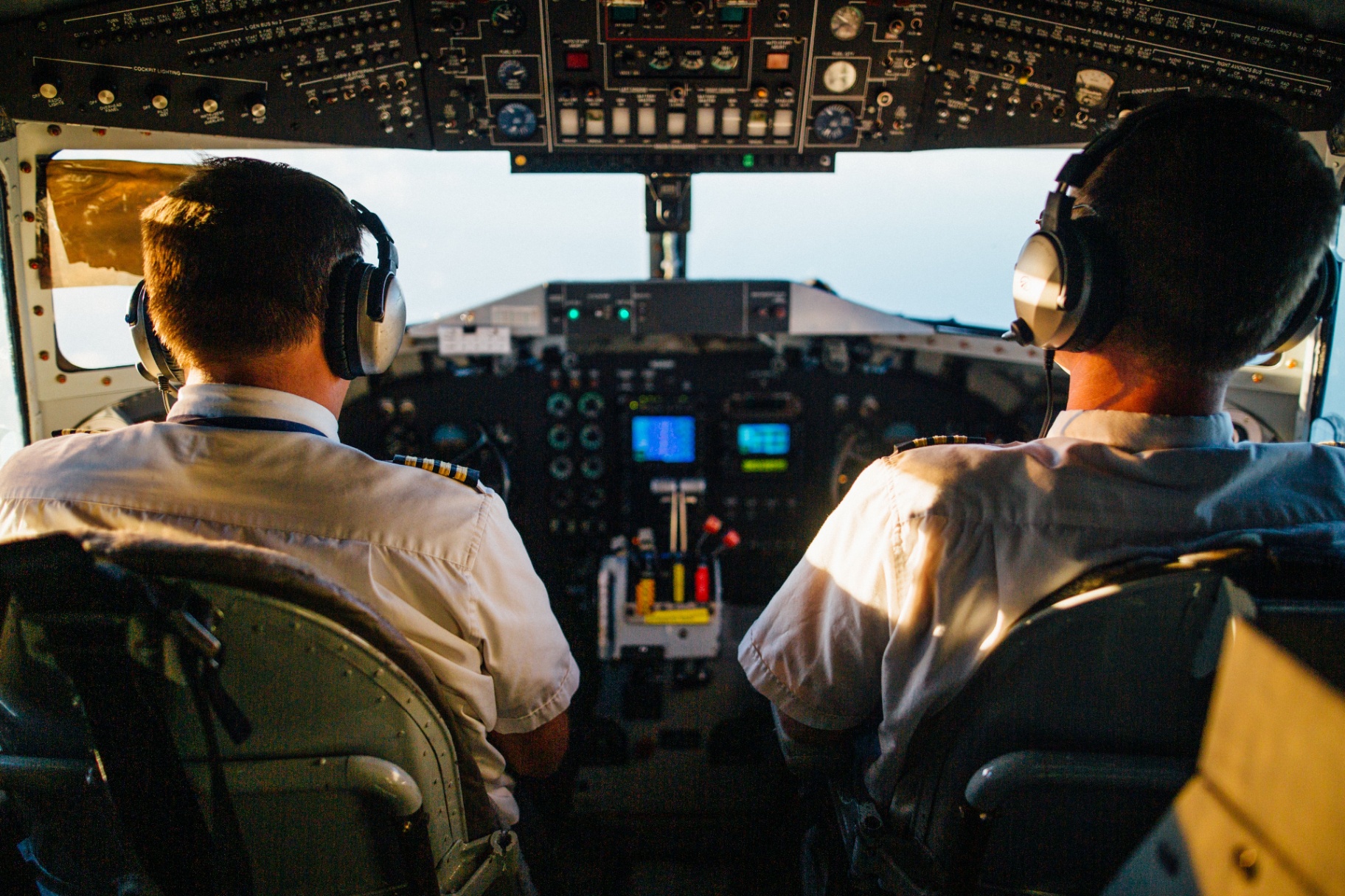 Two pilots at the flight controls seen from behind, focused on the instrument panel and horizon ahead