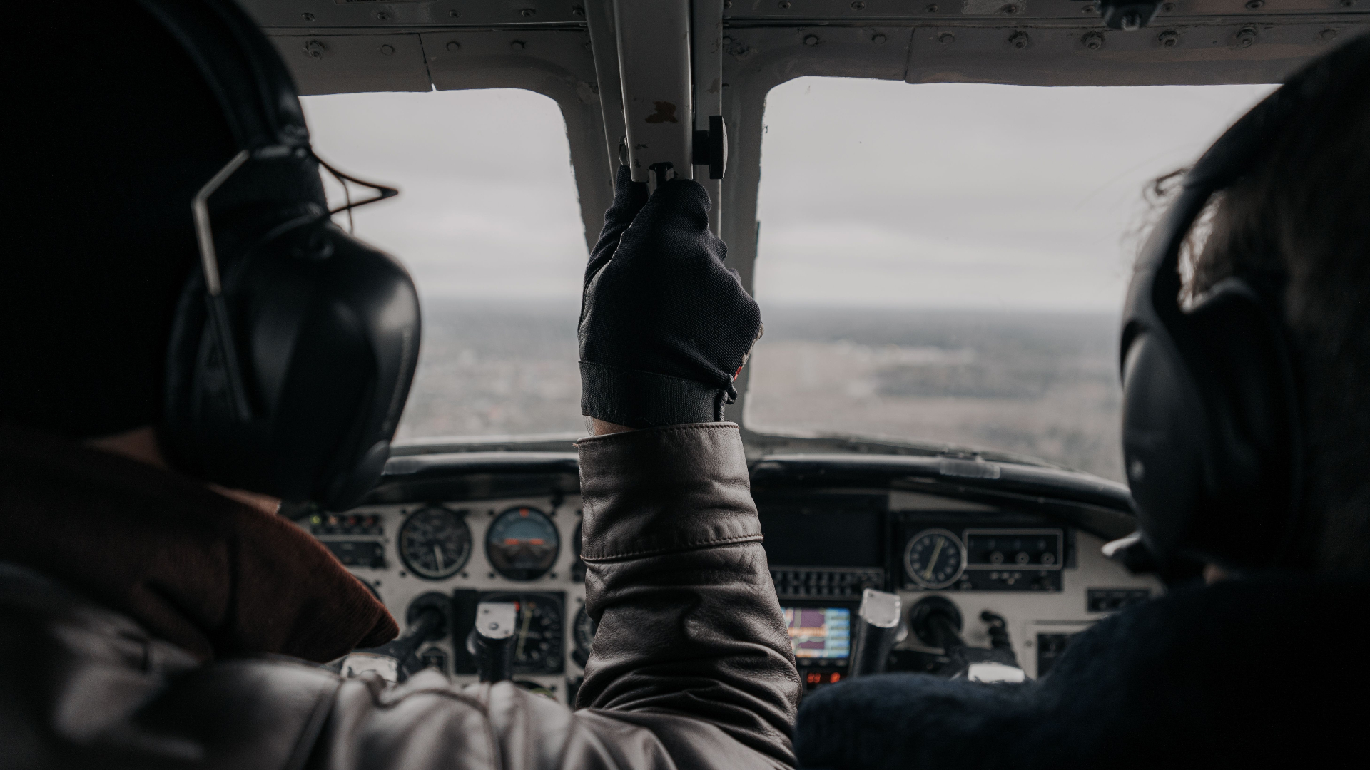 View from behind two pilots wearing headsets flying a small aircraft over an overcast landscape